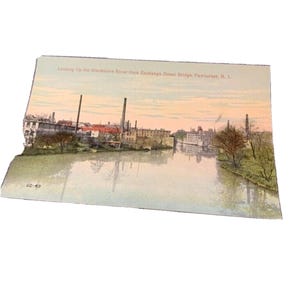 May include: A vintage postcard depicting the Blackstone River from Exchange Street Bridge, Pawtucket, R.I. The image shows a river scene with buildings and chimneys along the banks, under a pastel sky. The postcard is slightly angled.