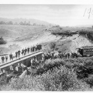 Può includere: Una fotografia in bianco e nero di un treno su un binario. Il treno è circondato da persone e l'anno 1906 è scritto nell'angolo in alto a destra dell'immagine.