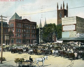 May include: A vintage postcard depicting a bustling public market in Albany, New York. The scene features a variety of horse-drawn carriages and wagons, as well as a large brick building with the text "Lyon Block" and "International Shirt Company" displayed on the facade. The postcard is dated "Oct. 11, 1906" and includes the handwritten inscription "Where is Harvey?"