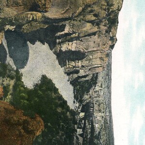 May include: Vintage postcard featuring a scenic landscape with a towering rock formation and a body of water. The image showcases a rugged cliff face with varying shades of brown and gray, contrasted by a light blue sky.