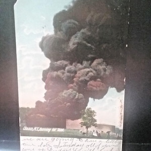 Op de afbeelding: Vintage ansichtkaart met de brandende olietank in Olean, NY. De afbeelding toont een grote wolk van donkere rook die de lucht in opstijgt. De ansichtkaart heeft handgeschreven tekst aan de onderkant, met de woorden "Olean, NY, Burning Oil Tank" bovenaan gedrukt.