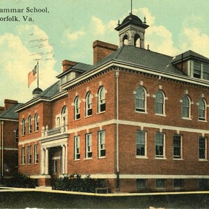 Op de afbeelding: Oude ansichtkaart met de High and Grammar School in South Norfolk, Va. Het rode bakstenen gebouw heeft boogvormige ramen, een koepel en een Amerikaanse vlag. De lucht is lichtblauw met wolken.