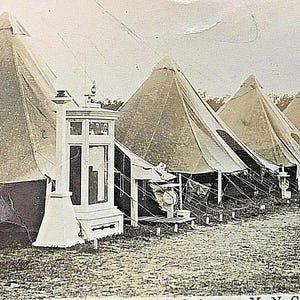 Pode incluir: Fotografia a preto e branco de um acampamento militar. Várias tendas grandes em forma de cone estão alinhadas. Uma estrutura branca com um topo decorativo está em frente às tendas. A imagem está ligeiramente desbotada.