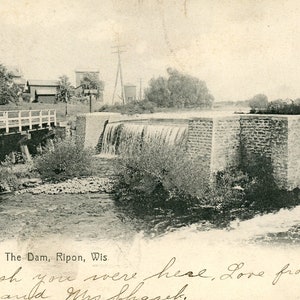 Pode incluir: Um cartão postal vintage em preto e branco mostrando uma barragem com uma ponte de madeira sobre um rio. O texto no cartão postal diz "The Dam, Ripon, Wis. Wish you were here. Love from Jack and Mrs. Shoresh."