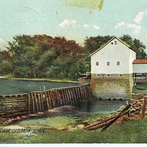 Puede incluir: Una postal antigua que representa un edificio de madera blanco con una base de piedra, situado junto a un pequeño lago. El edificio es una presa de molino en Hazelton, Iowa. La postal tiene un aspecto antiguo y descolorido.
