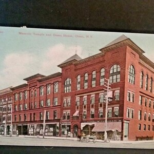 Op de afbeelding: Een vintage ansichtkaart met de Masonic Temple en Olean House in Olean, NY. De afbeelding toont een groot, meerverdiepingen tellend bakstenen gebouw met boogvensters en sierlijke details. Een paard en wagen is zichtbaar op straat.