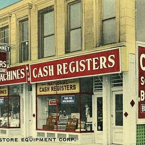 May include: Vintage storefront with signs reading "CASH REGISTERS" and "SLICING MACHINES." The business, "BURL & KENNEY," also offers scales and repair services. The building has multiple windows and a classic architectural style.