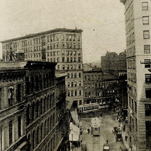 May include: An elevated view of a city street scene from the early 20th century. Several multi-story buildings line the street, with a trolley car and pedestrians visible. The image is in black and white.