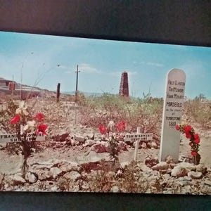 May include: Vintage postcard featuring a desert landscape with a tombstone and grave markers. The tombstone reads "Billy Clinton, Tom McLowery, Frank McLowery, Murdered, Tombstone 1881." Red and white flowers adorn the graves.