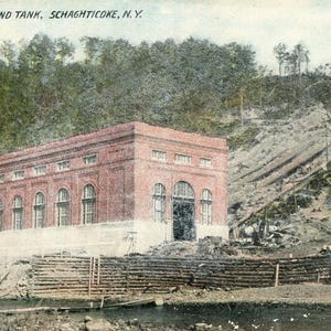 May include: A vintage postcard depicting a brick power house and tank in Schaghticoke, New York. The building is surrounded by a wooden fence and a river.