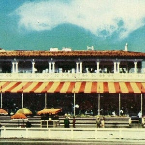 Puede incluir: Imagen de postal vintage de un edificio con un toldo a rayas rojas y amarillas. El edificio tiene una torre blanca con una bandera y un balcón. Se ven varios parasoles frente al edificio.