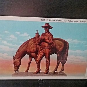Op de afbeelding: Een vintage ansichtkaart met een bronzen beeld van een cowboy en zijn paard. De cowboy staat naast het paard en houdt de teugels vast. Het paard is gebogen en graast. De achtergrond toont een blauwe lucht met wolken. Tekst bovenaan luidt: "5211—A Range Rider of the Yellowstone, Billings, Mont."