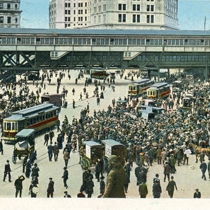 May include: A vintage black and white postcard showing a busy city street scene with a large elevated railway structure overhead. The street is packed with people, some walking, some standing, and some riding on trams. There are several buildings in the background, including a tall building with a clock tower.