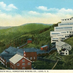 Può includere: Una cartolina d'epoca che raffigura un grande complesso industriale con più edifici, tra cui un edificio bianco a più piani con molte finestre e diversi edifici rossi più piccoli. Il complesso è situato su una collina con alberi verdi e vegetazione. La cartolina recita "The New South Mill, Homestake Mining Co., Lead, S.D."