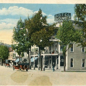 May include: A vintage postcard depicting a hotel called "Hotel DeCumber" with a large sign above the entrance. The hotel is a two-story building with a porch and several windows. There are trees in front of the hotel and a few cars parked in the street.