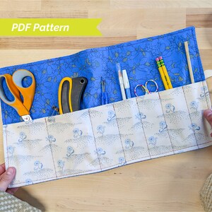 May include: A blue and white fabric roll-up case with a floral pattern. The case is open and contains various tools, including scissors, a craft knife, pencils, and a wooden dowel.