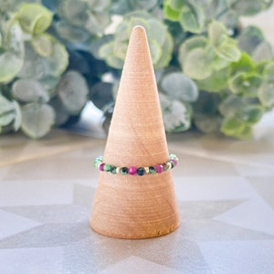 May include: A green, pink, and gold beaded ring on a wooden ring holder. The ring is made of small, round beads.
