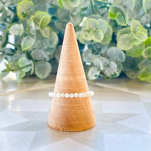 May include: A white beaded ring on a wooden display stand. The ring is made of small, round beads that are strung together to form a simple band.