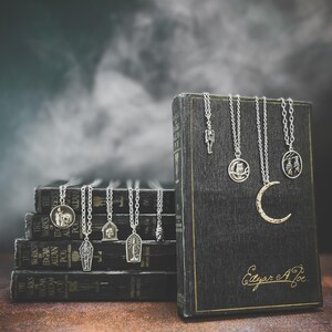 Puede incluir: Una colecci&oacute;n de collares plateados con colgantes de tem&aacute;tica g&oacute;tica, incluyendo un esqueleto y una luna creciente, se exhiben sobre una pila de libros antiguos. Los libros son oscuros con letras doradas, y la imagen tiene un fondo atmosf&eacute;rico.