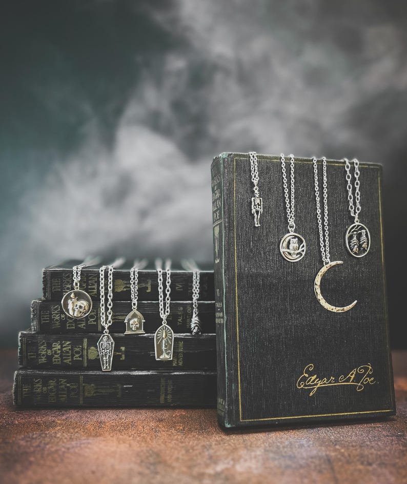 Puede incluir: Una colecci&oacute;n de collares plateados con colgantes de tem&aacute;tica g&oacute;tica, incluyendo un esqueleto y una luna creciente, se exhiben sobre una pila de libros antiguos. Los libros son oscuros con letras doradas, y la imagen tiene un fondo atmosf&eacute;rico.
