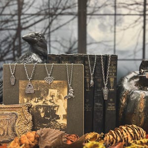 Puede incluir: Una exhibici&oacute;n tem&aacute;tica de Halloween con collares plateados con colgantes en forma de ata&uacute;d, murci&eacute;lago y telara&ntilde;a. Los collares se exhiben con libros antiguos, una estatua de cuervo y una calabaza decorativa.