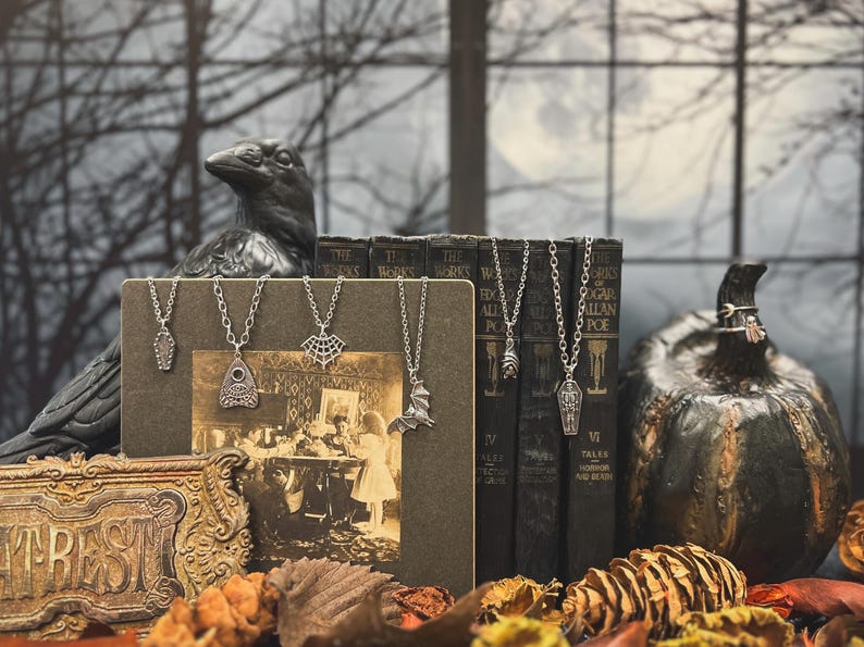 Puede incluir: Una exhibici&oacute;n tem&aacute;tica de Halloween con collares plateados con colgantes en forma de ata&uacute;d, murci&eacute;lago y telara&ntilde;a. Los collares se exhiben con libros antiguos, una estatua de cuervo y una calabaza decorativa.