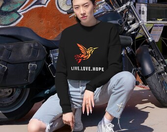 May include: A woman wearing a black cropped sweatshirt with a colorful hummingbird graphic and the text "LIVE.LOVE.HOPE". She is sitting in front of a motorcycle.