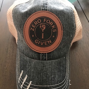 May include: A distressed charcoal gray baseball cap with a tan mesh back. The front features a circular brown leather patch with a black center displaying a cartoon character and the text "ZERO FORKS GIVEN" with two small stars.