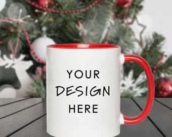 May include: White ceramic mug with a red interior and handle. The mug features the text "YOUR DESIGN HERE" in black. The background includes a blurred Christmas tree with ornaments and the website address TLLDESIGNS.COM.