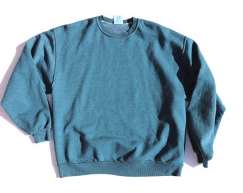 May include: A teal-colored crew neck sweatshirt. The long-sleeved top has a ribbed hem and cuffs. The garment is made of a soft material and is laid flat on a white surface. The sweatshirt is a casual style.