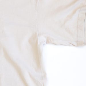 May include: A plain, light beige short-sleeved t-shirt featuring a standard crew neckline and visible stitching along the sleeve hem and side seam. The fabric has a smooth, solid-colored texture with no patterns, graphics, or additional embellishments.