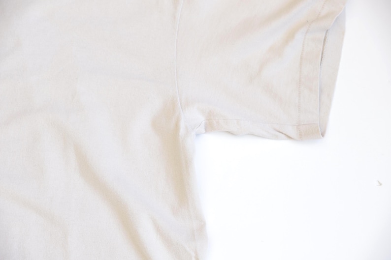 May include: A plain, light beige short-sleeved t-shirt featuring a standard crew neckline and visible stitching along the sleeve hem and side seam. The fabric has a smooth, solid-colored texture with no patterns, graphics, or additional embellishments.