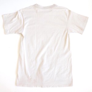 May include: A plain, solid light beige short-sleeve t-shirt shown from the back. The garment features a standard crew neckline, straight hemline, and simple construction with no visible patterns, graphics, or additional design elements.