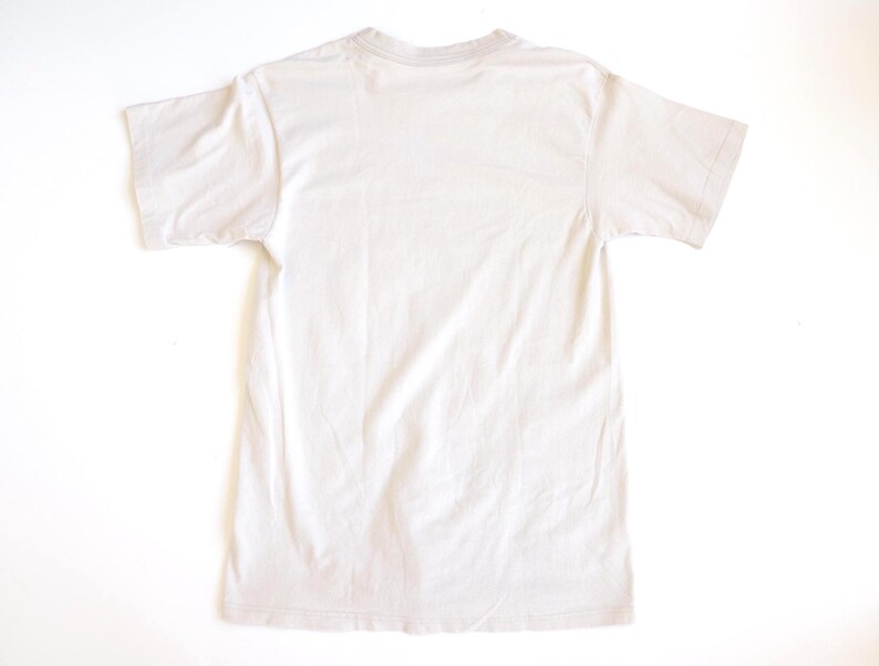May include: A plain, solid light beige short-sleeve t-shirt shown from the back. The garment features a standard crew neckline, straight hemline, and simple construction with no visible patterns, graphics, or additional design elements.