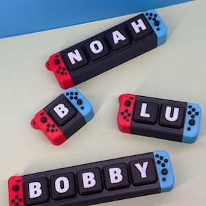 May include: Four custom name tags designed as game console controllers. Each tag has a black base with white letters spelling out names: NOAH, BOBBY, and LU. The tags have red and blue accents, resembling game console controllers.