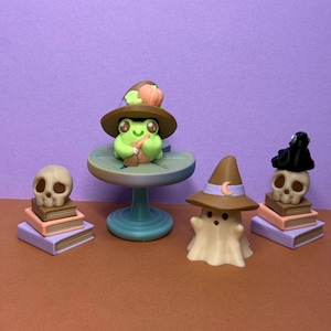 May include: A selection of Halloween figurines. A green frog wearing a witch's hat and holding a broom is on a small table. A ghost, a skull, and two stacks of books are also present. The figurines are in shades of green, brown, white, and purple.