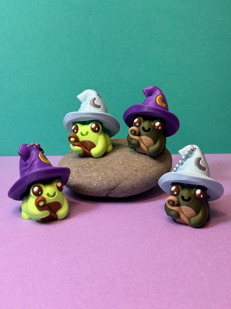 May include: Four whimsical frog figurines, each wearing a wizard hat with a crescent moon. The frogs are green and brown, and the hats are light blue and purple. They are posed on a gray stone against a teal background.