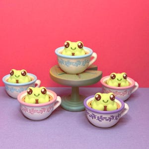 May include: Five frog figurines in teacups. The frogs are light green with large eyes and are nestled in pastel-coloured teacups with floral designs. One teacup sits on a small, round pedestal. The background is a vibrant pink and purple.
