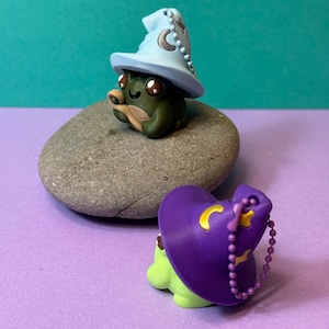 May include: Two whimsical frog figurines. One is dark green with a light blue wizard hat adorned with a crescent moon and stars. The other is light green with a purple wizard hat and a gold crescent moon. Both are on a gray stone.