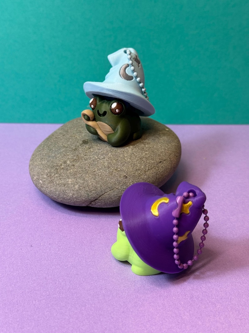 May include: Two whimsical frog figurines. One is dark green with a light blue wizard hat adorned with a crescent moon and stars. The other is light green with a purple wizard hat and a gold crescent moon. Both are on a gray stone.