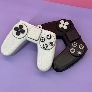 May include: Two game controllers, one white and one black, are shown against a purple backdrop. The controllers feature a classic design with directional pads, action buttons, and a square button. The white controller has a speckled texture.