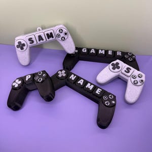 May include: A collection of black and white game controller ornaments are arranged on a purple surface. The controllers are inscribed with the words "SAM", "GAMER", "NAME", and "S". Each controller features buttons and a directional pad.