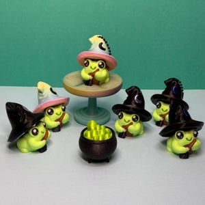 May include: A group of lime green frog figurines, each wearing a witch's hat. One frog sits on a small table, while others surround a miniature cauldron filled with glowing green liquid. Each frog holds a tiny broom.