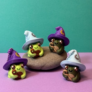May include: Four whimsical frog figurines, each wearing a miniature wizard hat. The frogs are green with brown accents, and the hats are light blue and purple with crescent moon details. They are arranged on a gray stone against a teal background.
