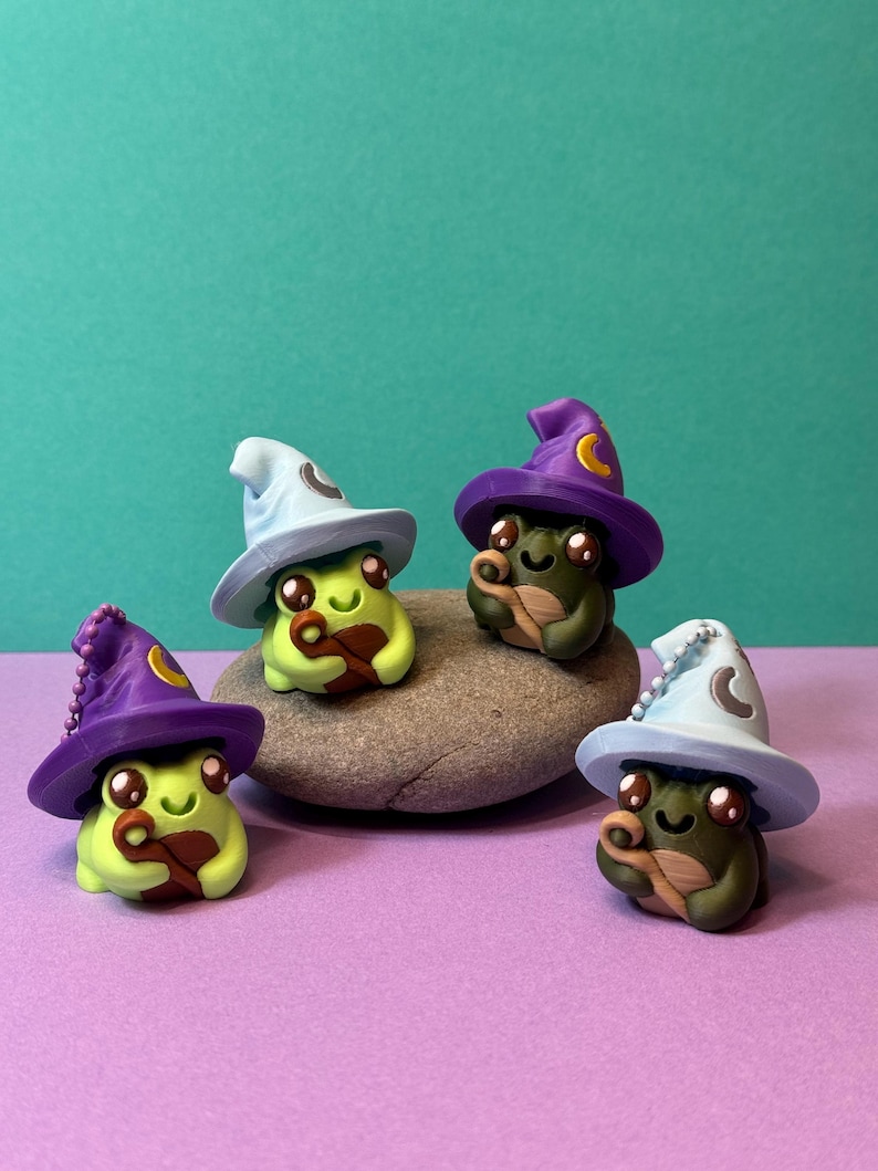 May include: Four whimsical frog figurines, each wearing a miniature wizard hat. The frogs are green with brown accents, and the hats are light blue and purple with crescent moon details. They are arranged on a gray stone against a teal background.