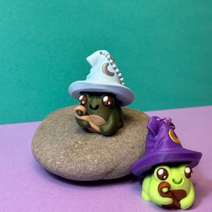 May include: Two whimsical frog figurines, one on a gray stone, each wearing a wizard hat. The first frog is green with a light blue hat and the second is lime green with a purple hat. Both have large brown eyes and are holding a brown object.