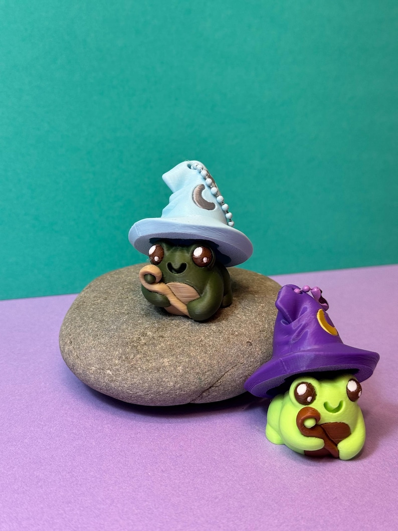 May include: Two whimsical frog figurines, one on a gray stone, each wearing a wizard hat. The first frog is green with a light blue hat and the second is lime green with a purple hat. Both have large brown eyes and are holding a brown object.