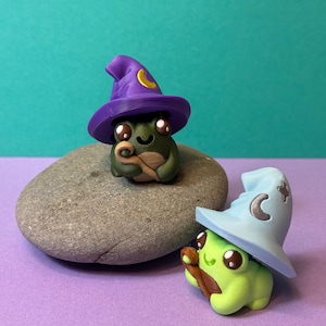May include: Two whimsical frog figurines, one dark green and one light green, each wearing a wizard hat. The dark green frog has a purple hat with a yellow crescent moon, while the light green frog has a light blue hat. They are on a gray stone.