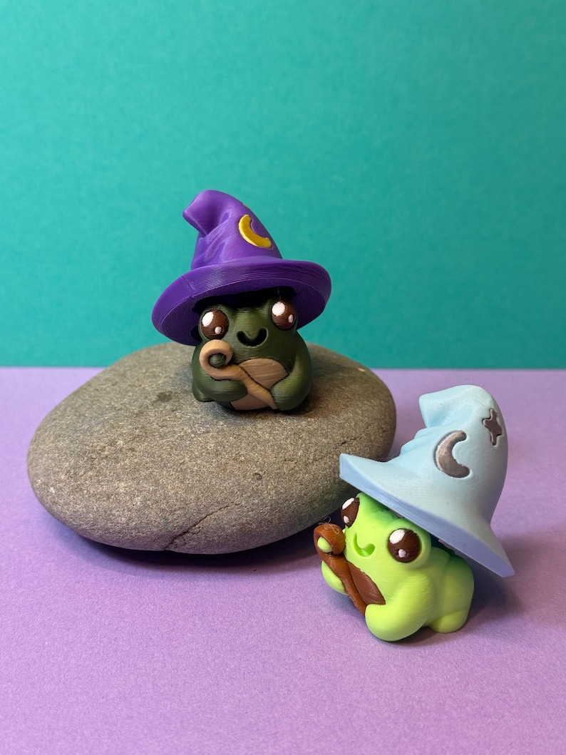 May include: Two whimsical frog figurines, one dark green and one light green, each wearing a wizard hat. The dark green frog has a purple hat with a yellow crescent moon, while the light green frog has a light blue hat. They are on a gray stone.