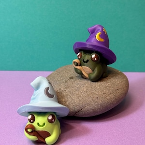 May include: Two whimsical frog figurines, one light green and one dark green, each wearing a wizard hat. The hats are light blue and purple, adorned with crescent moons and stars. The frogs are sitting on a gray stone against a teal and purple background.